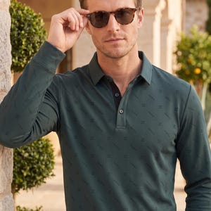 May include: A long-sleeve polo shirt in a dark green hue featuring a subtle, repeating diagonal pattern. The shirt has a classic collar, a buttoned placket with dark buttons, and ribbed cuffs. The fabric has a fine, textured, vertical ribbed finish.