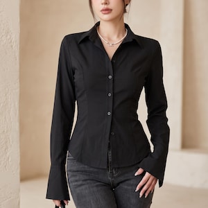 May include: A black long-sleeved button-down shirt with a pointed collar and flared cuffs, paired with dark grey denim jeans. The shirt features a fitted silhouette with vertical seams and a row of matching black buttons down the centre front.