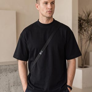 May include: A black short-sleeved t-shirt with a mock neck collar and a thin black crossbody strap featuring a small rectangular white label with black text. The shirt has a relaxed fit and is paired with dark navy blue trousers.