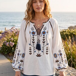 Op de afbeelding: Een witte blouse met lange mouwen, een split-halslijn met striksluiting en donkerblauwe kwastjes. Het kledingstuk is versierd met geborduurde bloemmotieven in blauwe, bruine en beige tinten op de voorzijde en de mouwen.