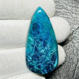 Puede incluir: Una gema en forma de lágrima con una superficie pulida y brillante. La piedra presenta una mezcla de tonos turquesa y azul oscuro, con intrincados patrones que se asemejan a vetas o grietas. La gema es sostenida por una mano enguantada.