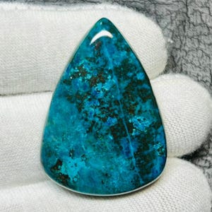 Puede incluir: Una gema pulida en forma de lágrima, con un vibrante tono turquesa, acentuada por inclusiones de color verde oscuro y negro. La piedra se encuentra sobre un fondo blanco, mostrando su belleza natural y patrones únicos.