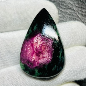 Pode incluir: Uma pedra preciosa polida em forma de gota. A pedra apresenta um centro rosa vibrante rodeado por tons verdes escuros e pretos. A gema é segurada em uma mão enluvada branca, mostrando sua beleza natural.