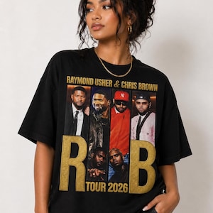 May include: Black t-shirt featuring a graphic of Raymond Usher & Chris Brown. The design includes images of the artists and the text "R&B TOUR 2026" in gold lettering. The shirt is a casual wear item.