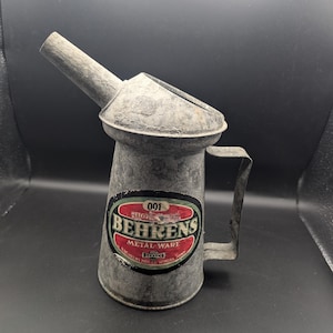 May include: Vintage Behrens metal ware oil can. The can is made of galvanized metal with a spout, handle, and a label with the text "Behrens Metal Ware". The label is red, white, and black. The can has a rustic, aged appearance.