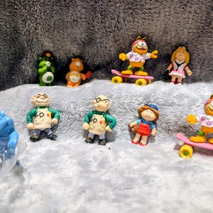 Puede incluir: Una colección de pequeñas figuras, incluyendo a Garfield en monopatín, Osos Amorosos y otros personajes. Las figuras son de colores brillantes y están sobre un fondo gris suave, creando una escena juguetona y nostálgica.