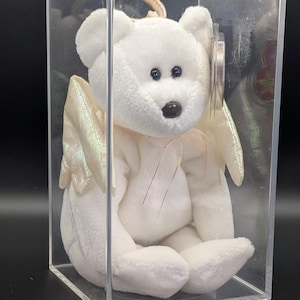 Peut inclure: Un ours en peluche blanc avec des ailes d'ange et un halo, présenté dans un boîtier en plastique transparent. L'ours a des yeux noirs et un nez brun. Les ailes ont un aspect irisé. Une étiquette est attachée à l'ours.