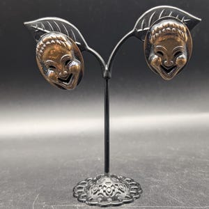 Puede incluir: Un par de pendientes vintage con caras de máscaras sonrientes. Los pendientes de color bronce tienen una pátina oscura, expuestos en un soporte negro. Las máscaras tienen rasgos faciales detallados y son de forma ovalada.