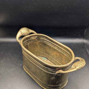 以下が含まれることがあります： 2つの装飾的なハンドルが付いた、真鍮製のアンティークな楕円形のプランター。 プランターは古びた外観で、経年劣化や使用の兆候が見られます。 ハンドルは装飾的で、エレガントさを添えています。 内側には変色が見られます。