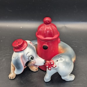 May include: A ceramic salt and pepper shaker set featuring a long-bodied dog wrapped around a bright red fire hydrant. The dog has grey and tan markings, a red hat on its head, and a small red bow with white polka dots on its tail end.