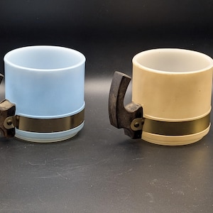 May include: Two vintage mugs, one light blue and one beige, each with a dark brown handle and a gold-colored band around the middle. The mugs are cylindrical with a slightly flared rim, set against a dark background.