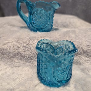 Op de afbeelding: Twee kleine, antieke, turquoise glazen kannen. De kannen hebben sierlijke, reliëfpatronen en gerimpelde randen. Eén kan heeft een handvat en een tuit, de andere is een kleine, open roomkan. Ze worden gepresenteerd op een zacht, wit oppervlak.