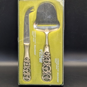 May include: A vintage cheese knife and slicer set in a green box. The silver-toned utensils have ornate handles and are labeled "ACANTHOS" and "Farmtinn". The set is made of hand-cast pewter and was made in Norway.