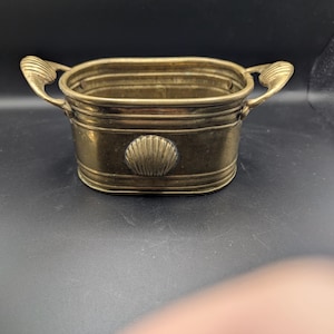 May include: A small, rectangular brass bucket with two ornate handles and a seashell design on the front. The bucket has a slightly aged appearance, suggesting it is vintage or antique. The overall design is simple yet elegant, suitable for decorative purposes.