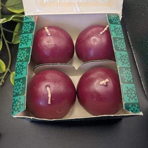May include: A box of four round, burgundy-colored candles. Each candle has a white wick. The box has a green and white snowflake design on the edges. The box is open, revealing the candles inside.