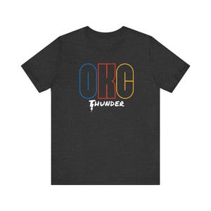 Op de afbeelding: Donkergrijs t-shirt met de tekst "OKC THUNDER" in een kleurrijk ontwerp. De letters "OKC" zijn omlijnd in blauw, rood en geel, met "THUNDER" in wit met een bliksem.