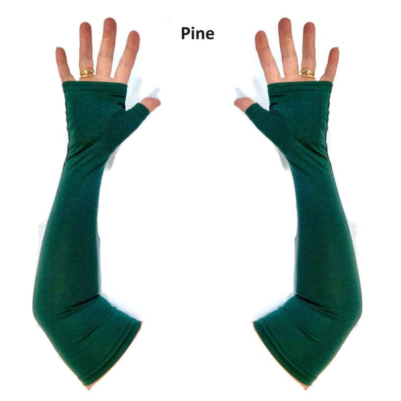 May include: A pair of long, fingerless arm warmers in a solid pine green colour. These sleeves extend from the knuckles to the upper forearm, featuring a thumb hole opening and a smooth, fitted fabric construction. The text Pine is visible.