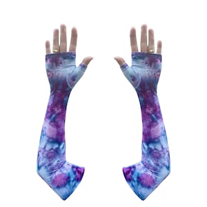 Puede incluir: Un par de mangas largas sin dedos con un estampado tie-dye en tonos morado, azul y blanco. Las mangas están diseñadas para cubrir los brazos y tienen un agujero para el pulgar para un ajuste seguro. La tela parece suave y elástica.