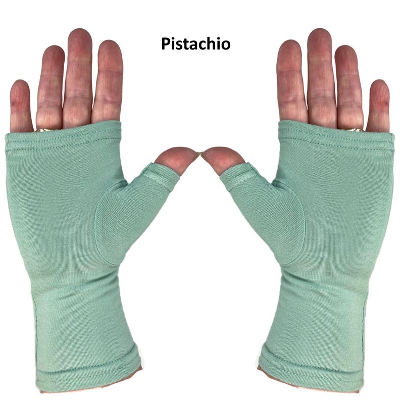 May include: A pair of fingerless gloves in a solid pistachio green color. The gloves feature a thumb hole and cover the palm and wrist area, with a stitched hem at the top and bottom edges. The text "Pistachio" is printed above the gloves.