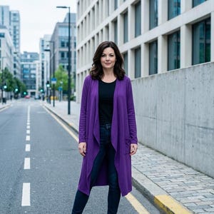 May include: A woman in a long, open-fronted purple cardigan, black top, and dark blue jeans, standing on a city street. She is also wearing black ankle boots. The background includes tall buildings and a cloudy sky.