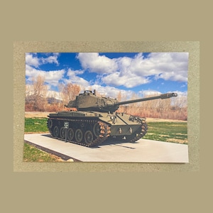 May include: An olive-green military tank with a long barrel, parked on a concrete slab. The tank is set against a backdrop of a blue sky with fluffy white clouds and a grassy field.