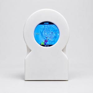 May include: A white, arch-shaped digital weather display device featuring a circular screen. The screen displays a blue radar map with the text HOME at the top and 15 mi at the bottom, showing various colored weather patterns over a geographic region.