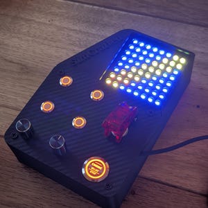 Puede incluir: Una caja de botones negra con textura de fibra de carbono y el texto SimCrafted, que cuenta con cuatro botones circulares naranjas iluminados, dos perillas giratorias plateadas, un interruptor rojo y una rejilla de luces LED azules y amarillas con un botón Engine Start Stop.