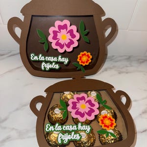 May include: Brown paper pot-shaped gift boxes with a clear window, decorated with pink and orange flowers and green leaves. The text "En la casa hay frijoles" is displayed on the front. One box is filled with gold-wrapped chocolates.