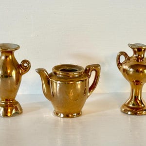 May include: A set of three miniature gold-toned ceramic decorative items consisting of two small vases with side handles and one central teapot with a spout and handle. Each piece features a reflective metallic finish and a smooth, polished surface.