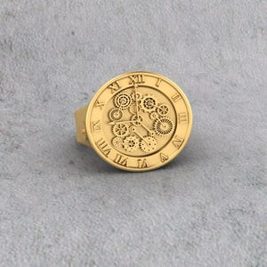 May include: Gold-colored ring with a clock face design. The ring features Roman numerals and intricate gear details within the clock face. The ring has a smooth, polished finish and a wide band.