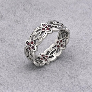 Op de afbeelding: Een zilverkleurige ring met een bloemmotief. De ring heeft kleine, ronde, rode edelstenen die zijn gezet in de bloemvormige elementen. De ring wordt getoond op een grijze ondergrond, wat de ingewikkelde details en het vakmanschap benadrukt.