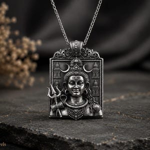 May include: A rectangular silver pendant on a chain featuring a detailed relief of a deity with a crescent moon, holding a trident. The design includes architectural elements on both sides of the figure and an ornate bail at the top.