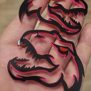 May include: Black and pink stylized dinosaur head cutouts with sharp teeth and red eyes. The cutouts have a glossy finish and are layered with a pink outline. The image shows three of these decorative cutouts.