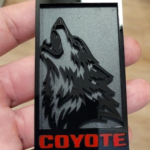 May include: Black rectangular emblem featuring a howling wolf silhouette. The wolf is set against a textured gray background, with the word "COYOTE" in red block letters at the bottom. The emblem is held in a hand.