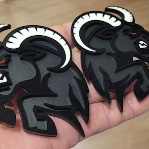 Puede incluir: Dos emblemas de cabeza de carnero negros con cuernos blancos y ojos rojos. Las cabezas de carnero están en una pose de salto, con un diseño detallado. Los emblemas probablemente están hechos de un material duradero.