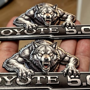 May include: Two metal emblems with a snarling wolf design. Each wolf is above a rectangular bar with the text "COYOTE 5.0L" in silver lettering. The emblems are detailed with shades of grey and black.