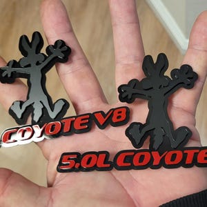 Puede incluir: Dos emblemas Coyote V8 negros y grises. Cada emblema presenta una figura de coyote de dibujos animados sobre el texto "5.0L COYOTE V8" en rojo y blanco. Los emblemas se sostienen en una mano.