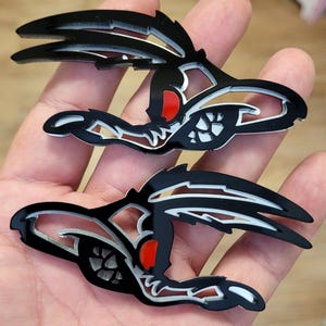 May include: Two black and white stylized decals featuring a bird-like design with a red accent. The decals have a glossy finish and are held in a person's hand. The design is intricate and detailed, with sharp angles and curves.