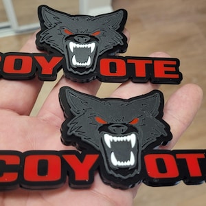 May include: Two black and red Coyote patches. Each patch features a snarling wolf head with red eyes above the word "COYOTE" in red letters. The patches are held in a hand.