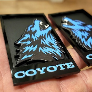 May include: Two black rectangular plaques with a blue and black howling coyote design. The word "COYOTE" is displayed in blue letters at the bottom of the plaque. The plaques are held in a hand.