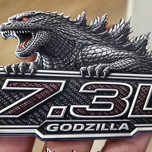 Puede incluir: Un emblema metálico detallado con Godzilla sobre una placa rectangular. La placa tiene el texto "7.3L GODZILLA" en un diseño de fibra de carbono con detalles en rojo. La figura de Godzilla es gris con ojos rojos.