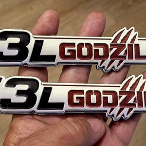 May include: Two identical rectangular automotive emblems featuring the text 7.3L GODZILLA in bold, raised lettering. The 7.3L is black with a red accent, while GODZILLA is dark red with a claw mark graphic integrated into the design on a white background.