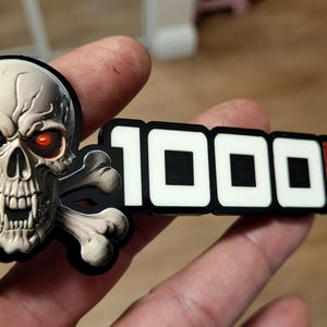 May include: A black and white emblem featuring a skull with red eyes and crossed bones. The text "1000HP" is displayed in white and red, indicating a high-performance theme. The emblem is held in a hand.
