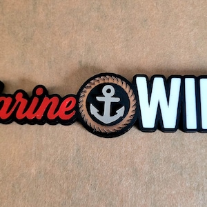 May include: A black and red Marine Wife emblem. The word "Marine" is in red script with a black outline. An anchor is in the center, and the word "WIFE" is in white block letters.