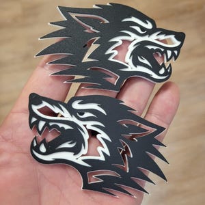 Op de afbeelding: Twee zwart-witte wolfskopuitsparingen. De wolfskoppen zijn in profiel, met scherpe tanden en een felle uitdrukking. De uitsparingen worden in een hand gehouden en de achtergrond is licht van kleur.