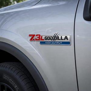 May include: A silver vehicle with a black tire and a chrome emblem. The emblem reads "7.3L GODZILLA HIGH OUTPUT" in red, white, and blue. The vehicle's body is a light silver color.
