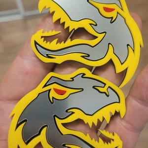 Può includere: Due adesivi a forma di testa di dinosauro. Ogni adesivo presenta una testa color argento con contorni gialli e denti affilati. Gli occhi sono rossi. Gli adesivi sono tenuti in una mano.
