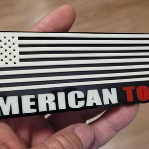 May include: A rectangular black and white emblem featuring the American flag design with the text "AMERICAN TOUGH" in white and red. The emblem is held in a hand.