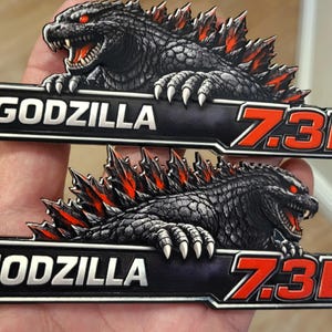 May include: Two black and gray Godzilla emblem badges with red accents. Each badge features the word "GODZILLA" and "7.3L" in white and red lettering. The badges depict a detailed Godzilla design.