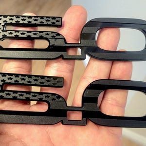 May include: Two black 5.0 emblems. The '5' features a star pattern, while the '0' has horizontal lines. These emblems are likely for a vehicle, possibly a Ford Mustang, and are designed to be affixed to the exterior.